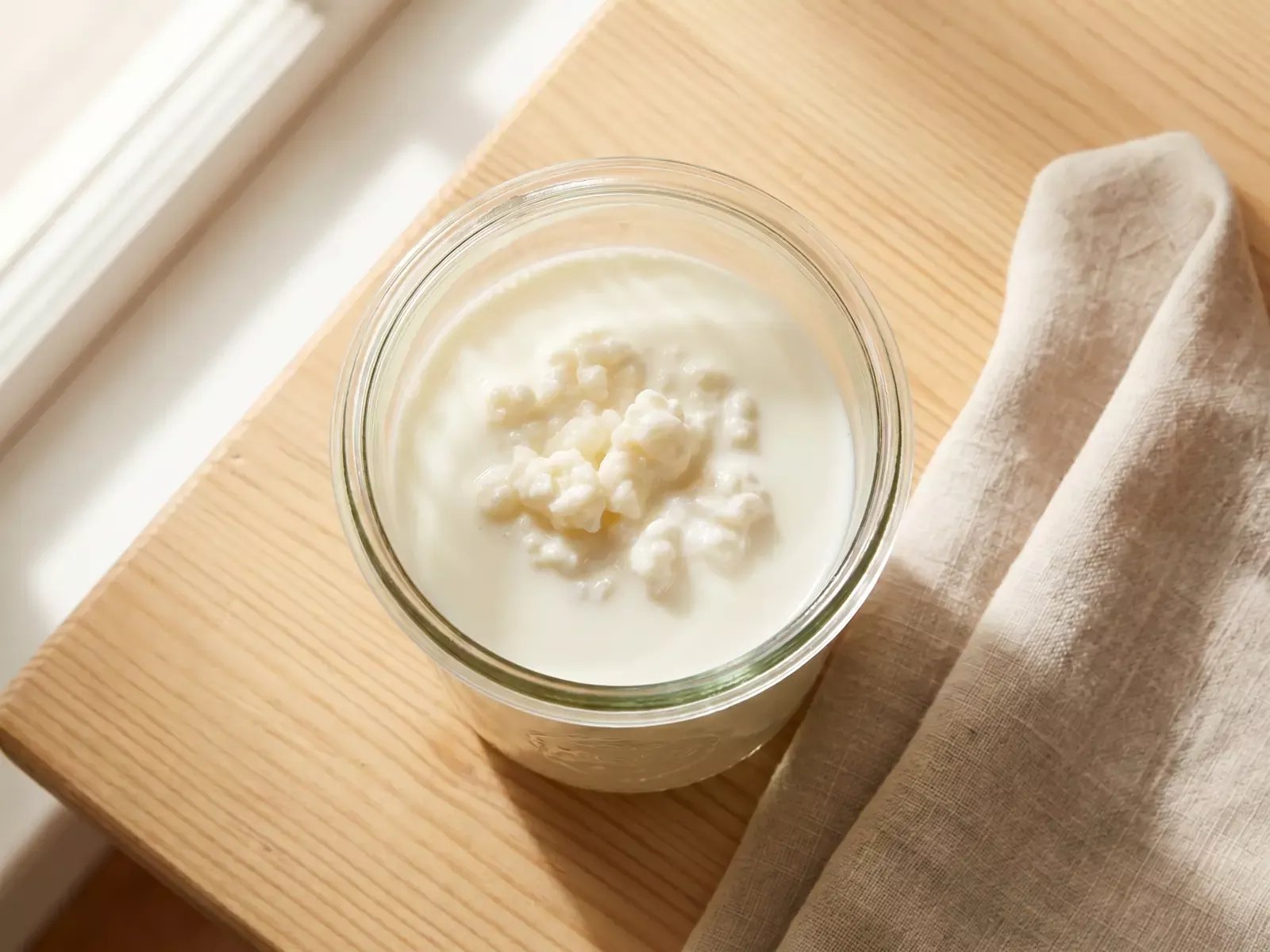 Glazen pot met melkkefir die fermenteert naast een linnen doek op een houten ondergrond.