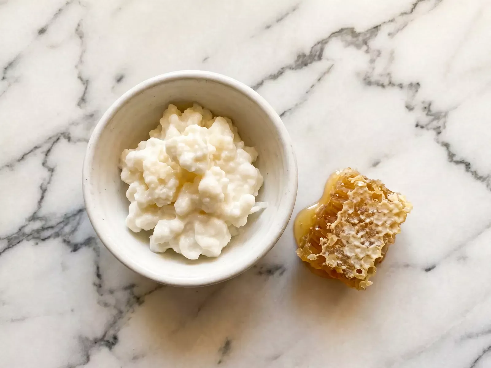 Kom met natuurlijke melkkefir naast honingraat op licht marmer, warm van bovenaf belicht.