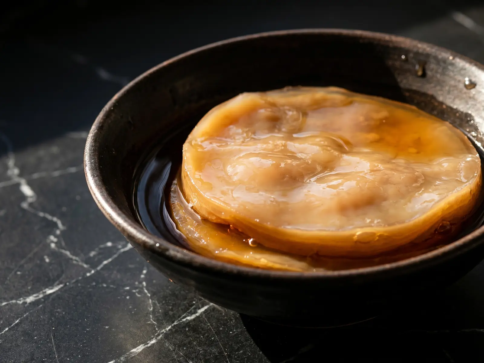 kombucha scoby Cuenco met donkere thee en een beige bruine SCOBY op mat zwart marmer in dramatisch daglicht.