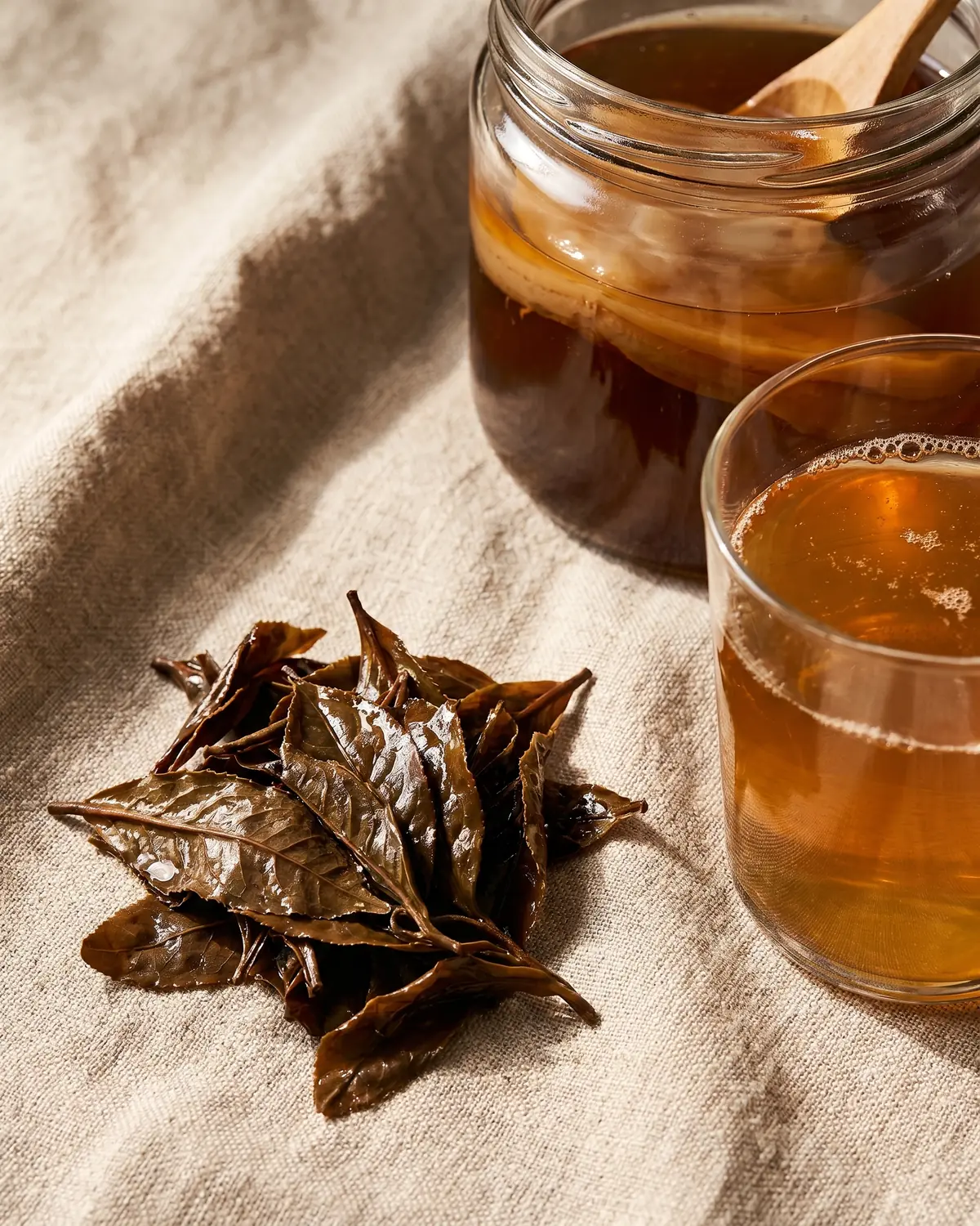 Vochtige theebladeren naast een glas kombucha op ruwe linnen stof met zachte natuurlijke kleuren.