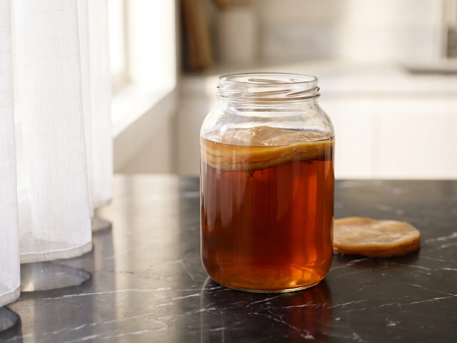Glazen pot met SCOBY en diepe amberkleurige thee op mat zwart marmer met witte gordijnreflectie.