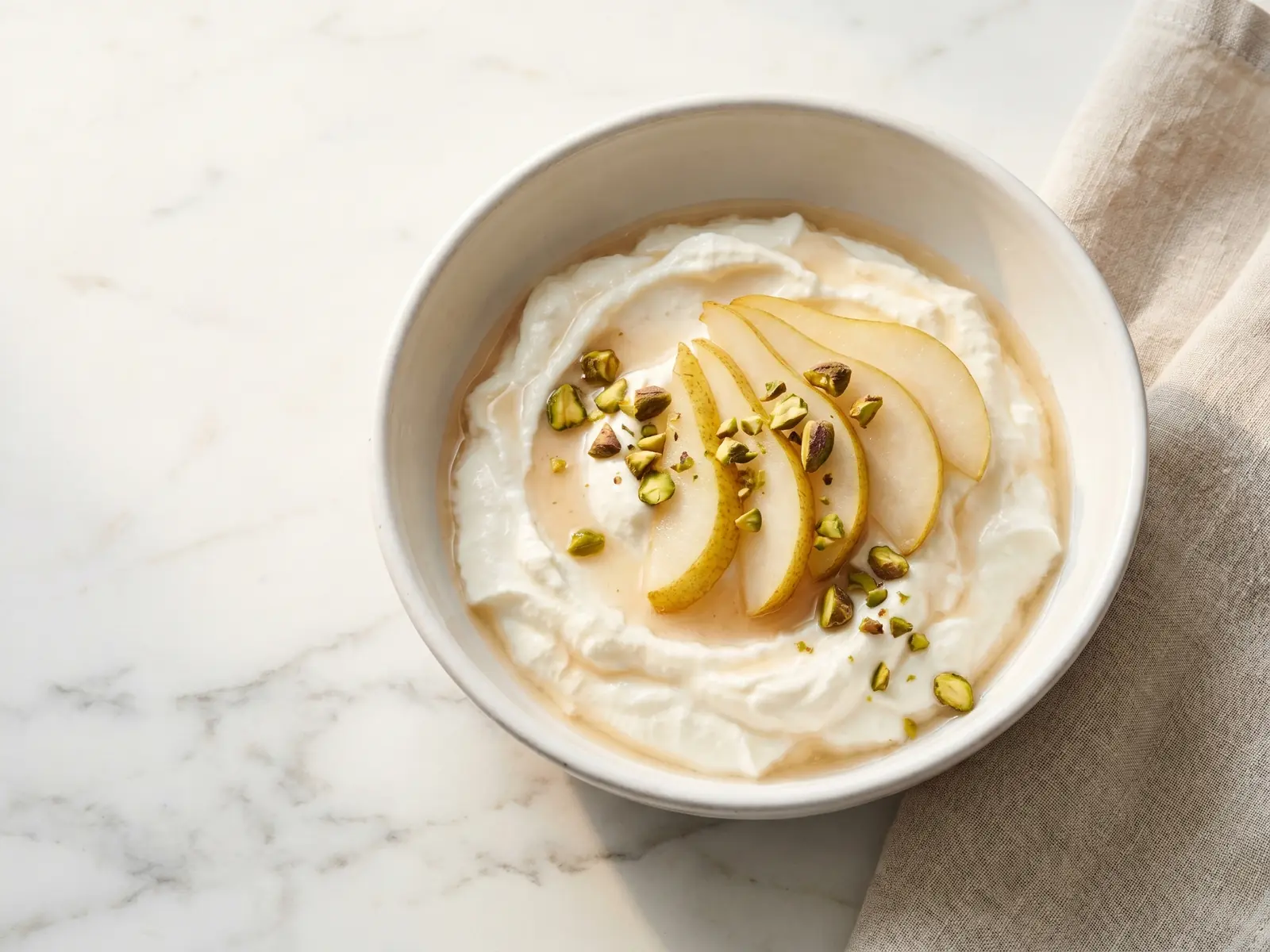Romige yoghurt in een witte keramische kom met plakjes peer en pistachenoten op geaderd marmer.